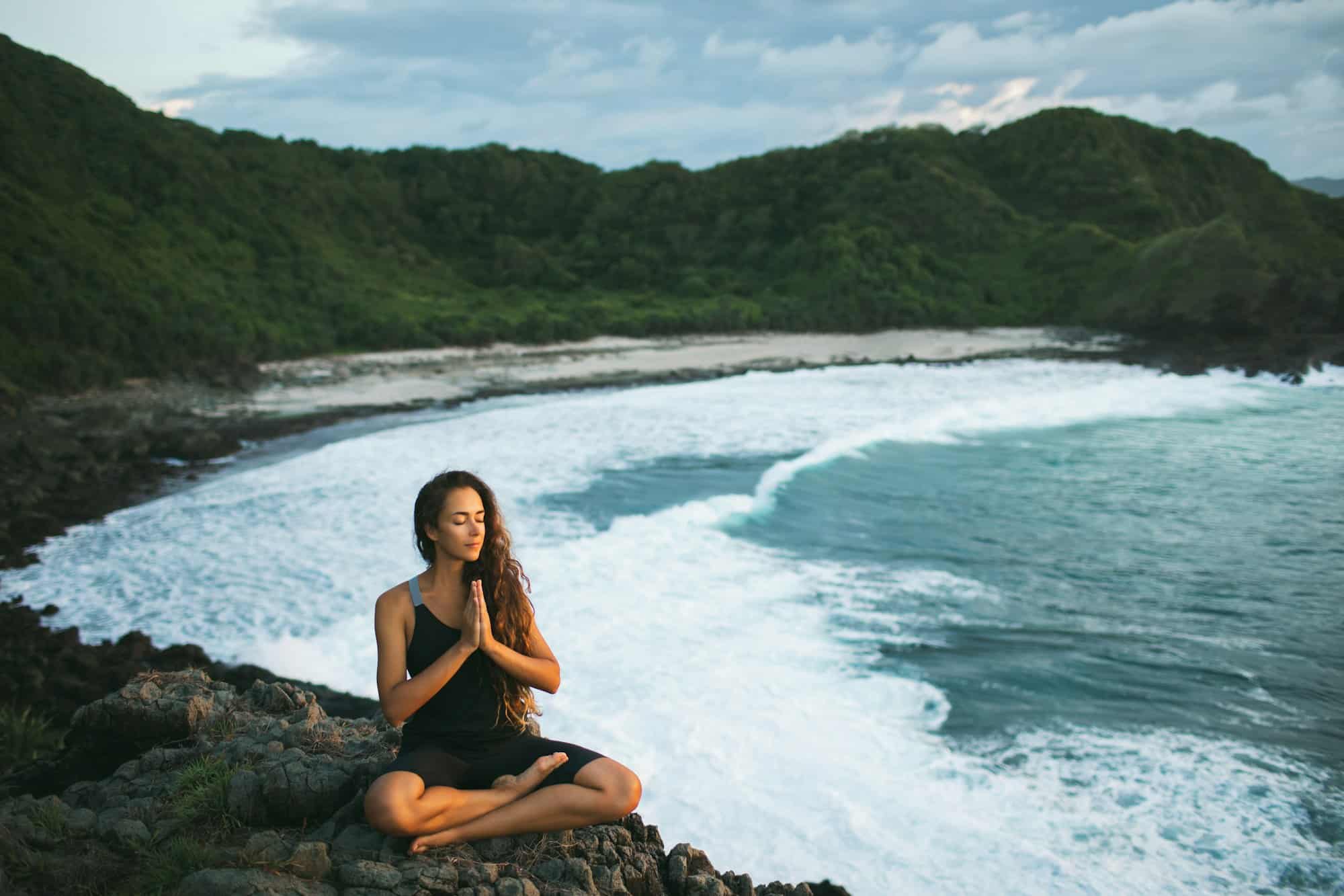 Young woman praying and meditating alone at sunset with beautiful ocean and mountain view. Self care