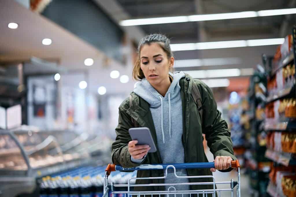 Young woman using shopping app on mobile phone while buying in supermarket.